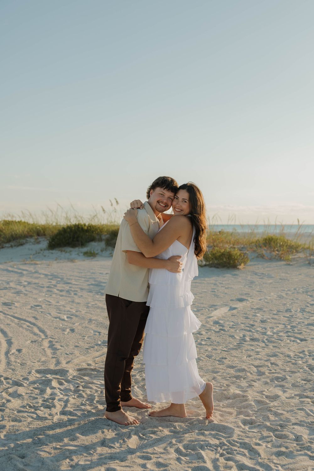 A couple embraces barefoot on a sandy beach at sunset, the woman in a white dress smiling at the camera.