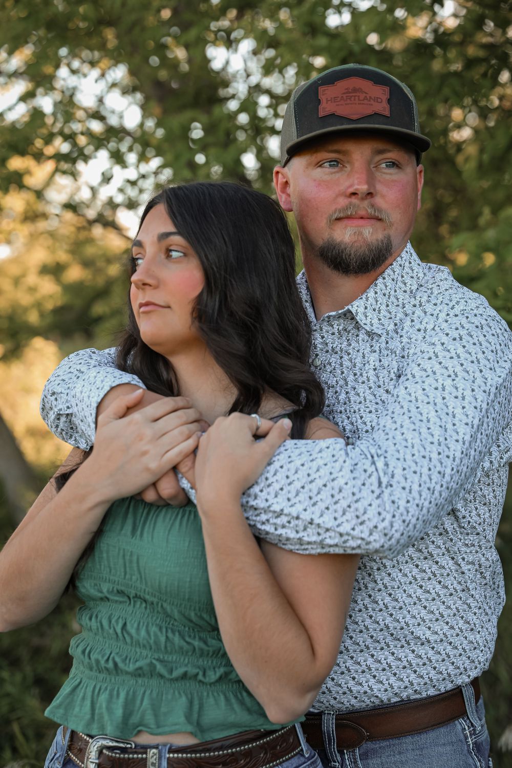 Couple embracing outdoors, man in cap and patterned shirt holding woman in green top, with lush green trees behind them.