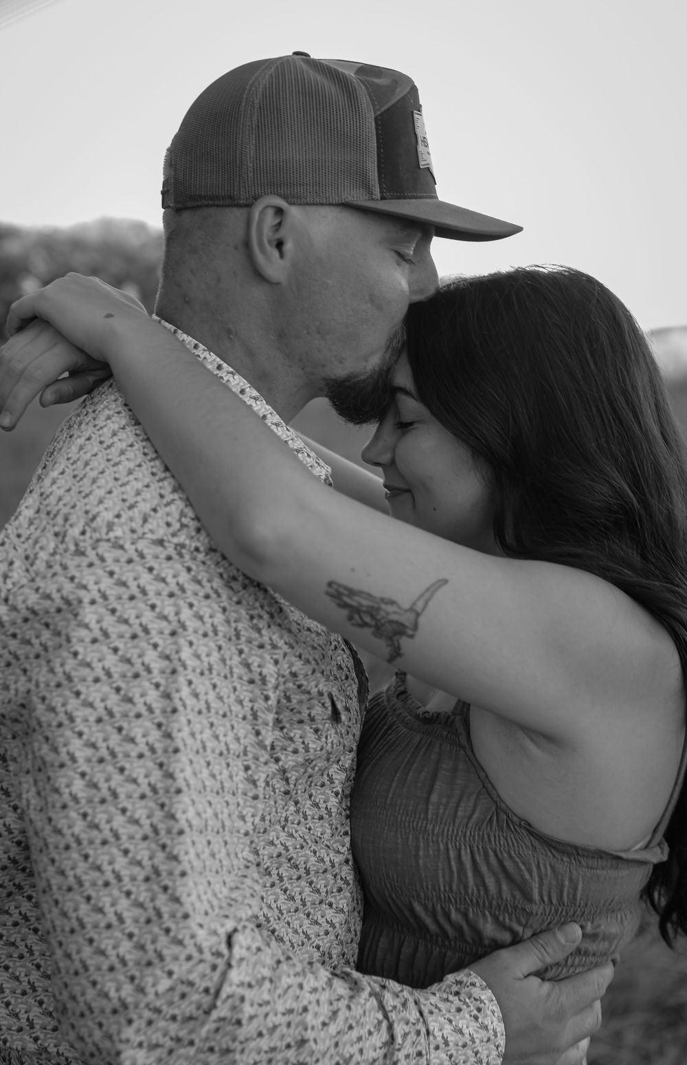 Black and white photo of a couple embracing and kissing outdoors, man in cap and patterned shirt, woman with arm tattoo.