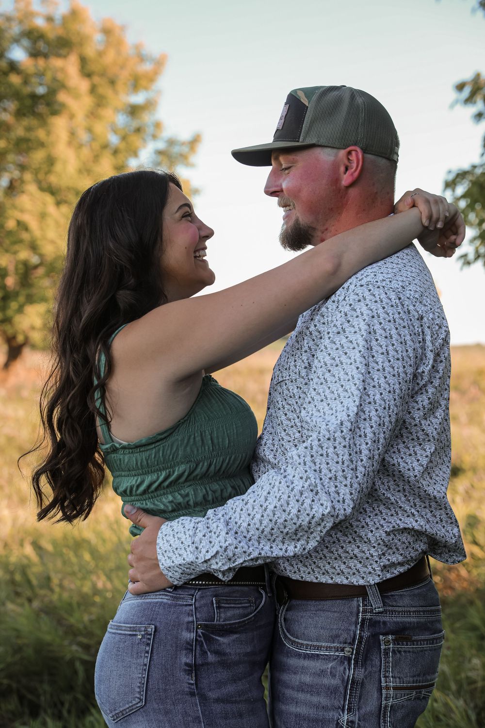 A smiling couple embraces outdoors, the woman in a green top and jeans, the man in a patterned shirt and cap.