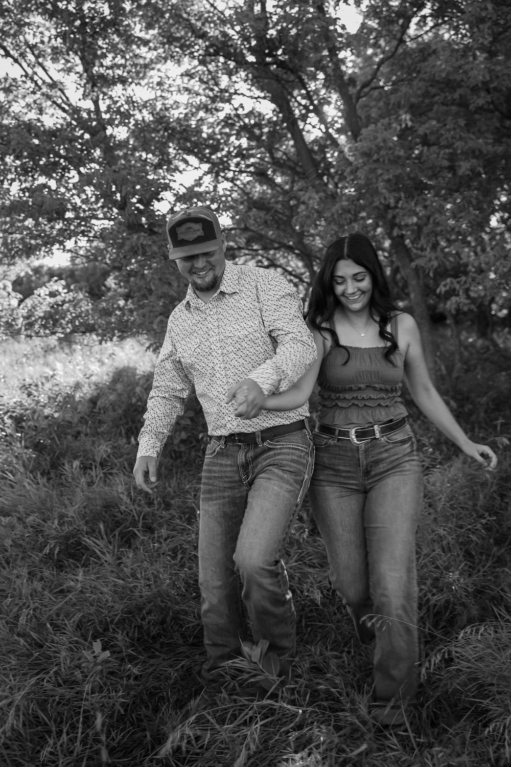 Black and white photo of a smiling couple walking together through a grassy field with trees in the background.