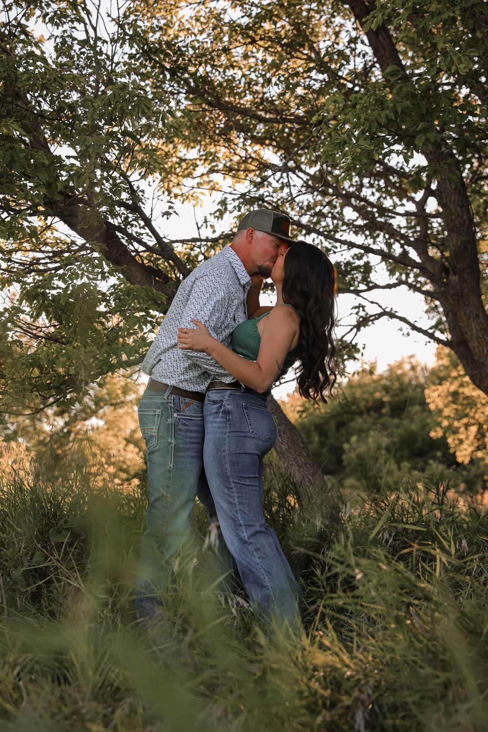 Couple kissing romantically under a large tree at golden hour, wearing jeans and casual western attire in a lush outdoor setting.
