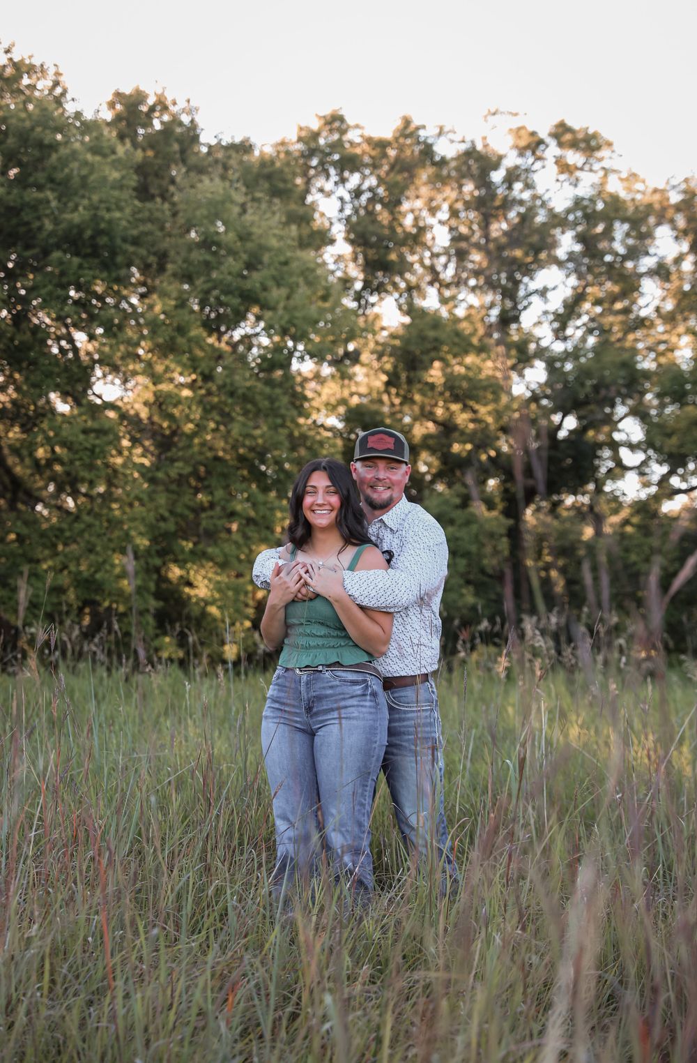 Couple embracing in tall grass field with lush green trees in golden hour light, wearing jeans and casual tops.