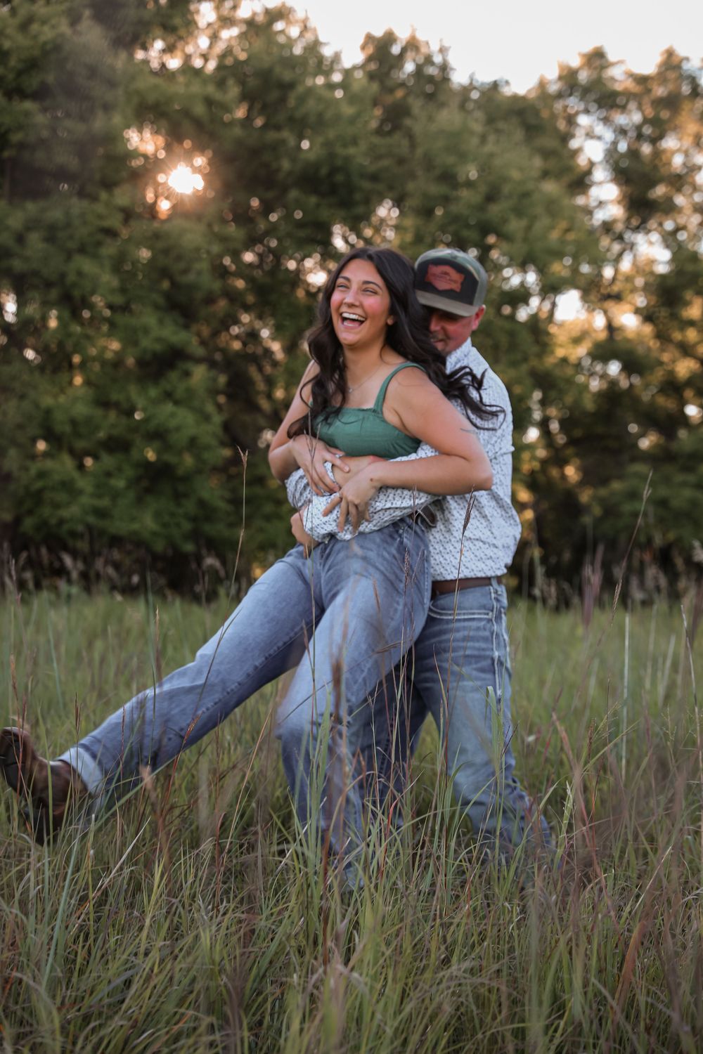 Couple laughing in tall grass field at golden hour, woman lifted by man in cap and western shirt.