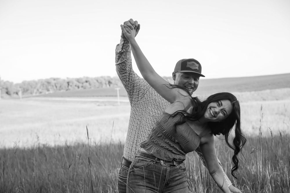 Black and white photo of a happy couple dancing together in a field, man in cap twirling woman who laughs joyfully.