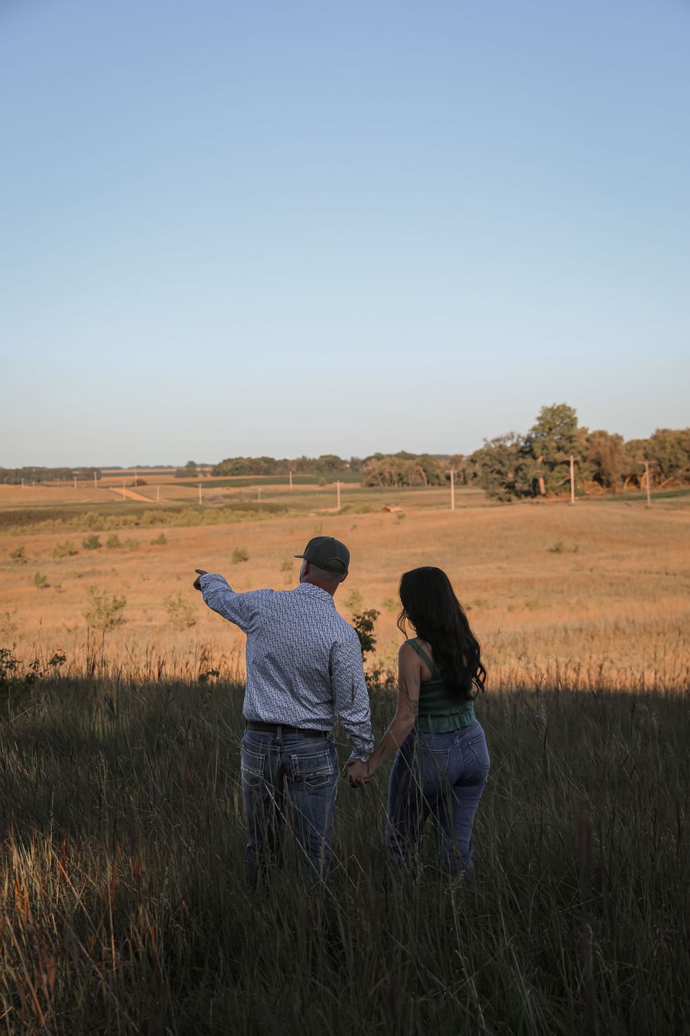 Couple standing in a golden field at sunset, man pointing toward the horizon while holding hands with woman.