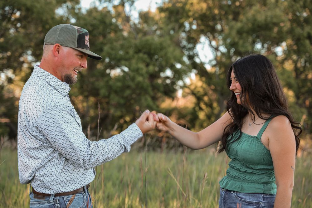 A smiling couple holds hands in a green meadow, man in plaid shirt and cap, woman in green top, during golden hour.