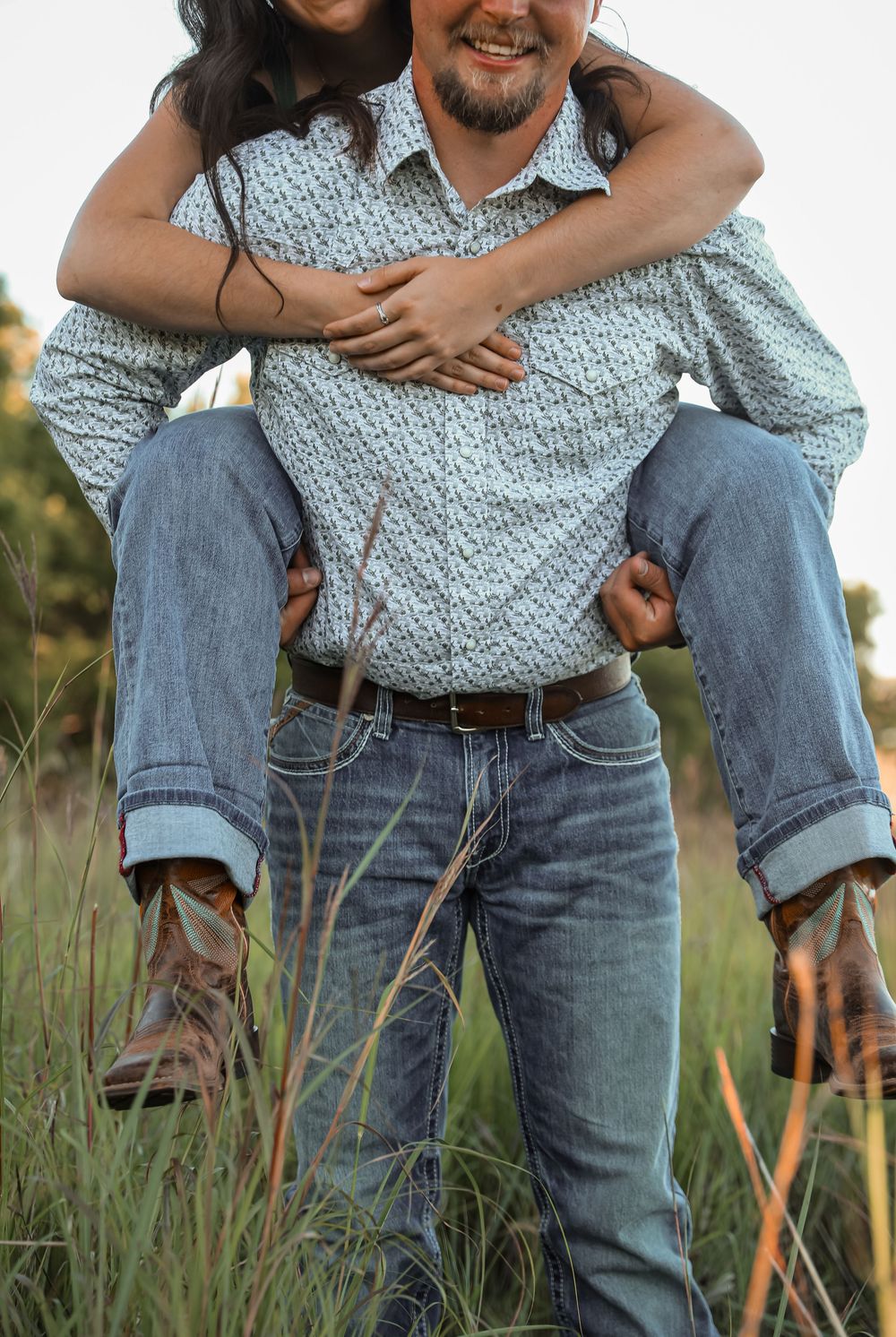 A man gives a woman a piggyback ride through a grassy field, both smiling and wearing jeans and boots.