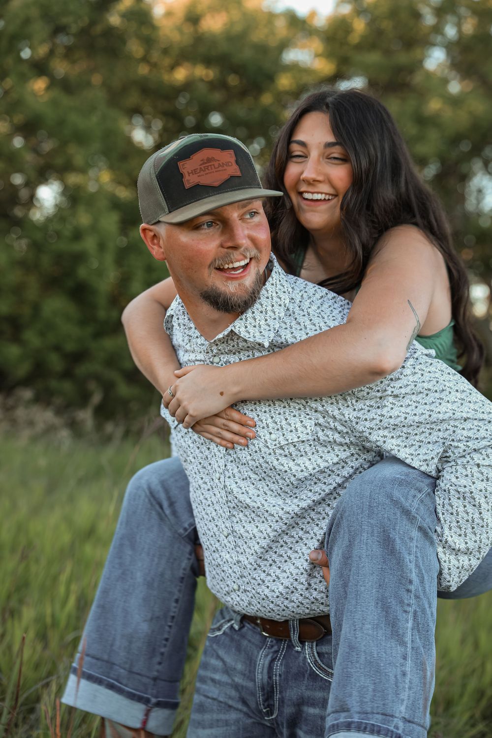 Happy couple laughing outdoors, man in trucker hat giving woman a piggyback ride.
