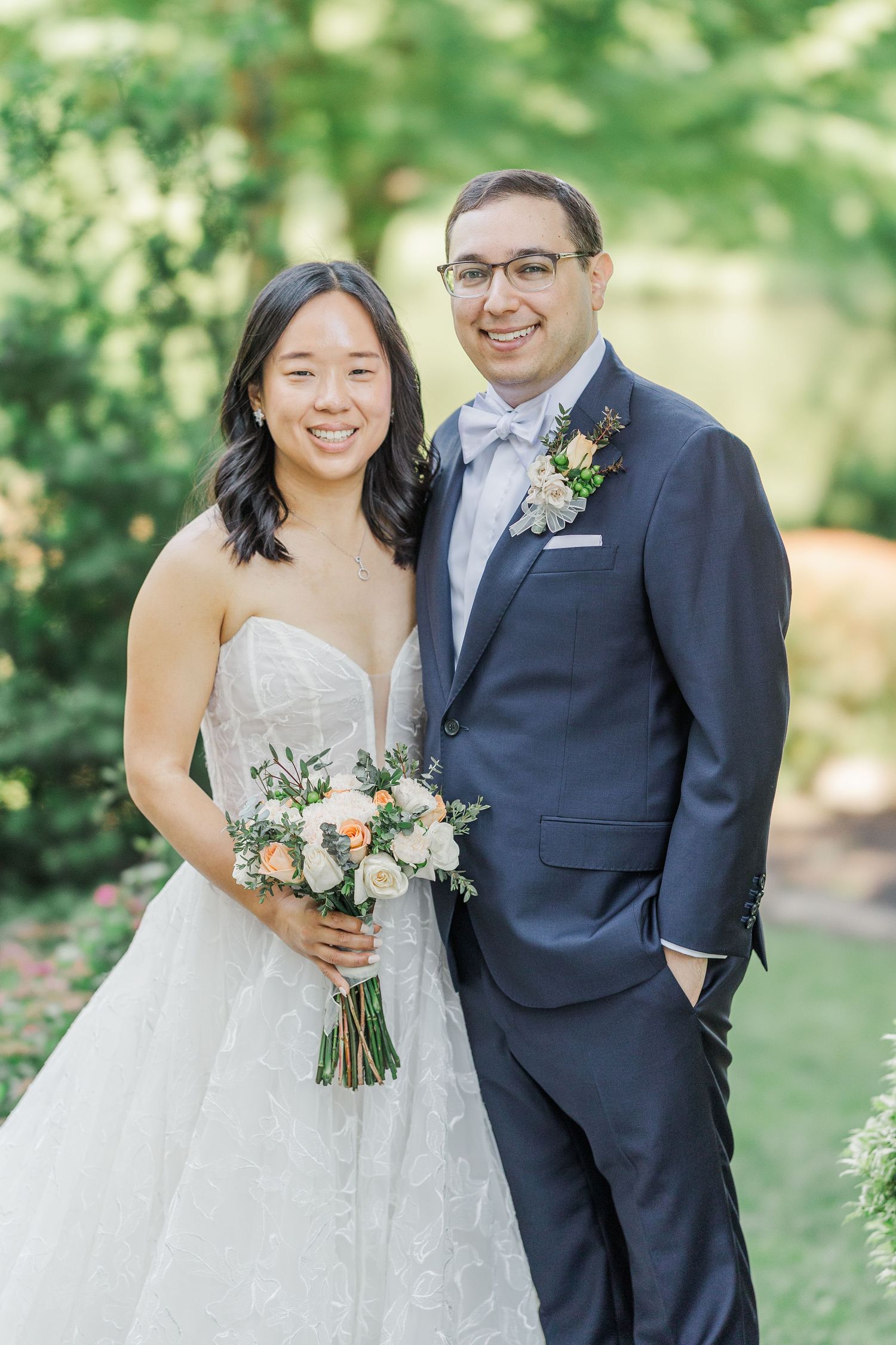 Bride in strapless lace gown holding peach bouquet stands with groom in navy suit and bow tie outdoors.