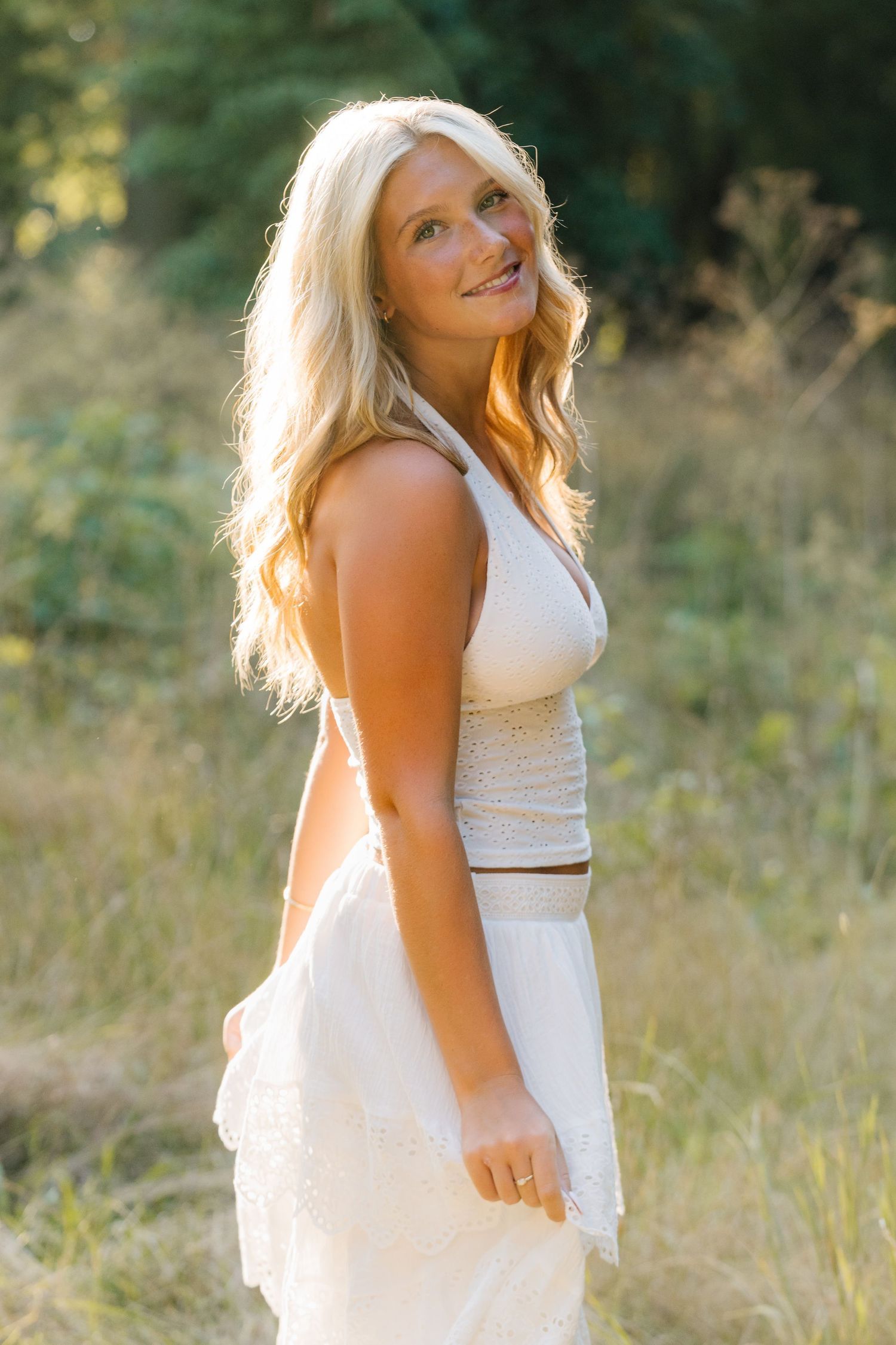Smiling blonde woman in white dress standing in sunlit field with green trees in background.