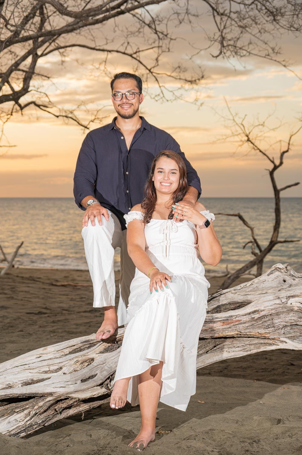 Couple posing on driftwood at beach sunset, woman in white dress, man in navy shirt and white pants.