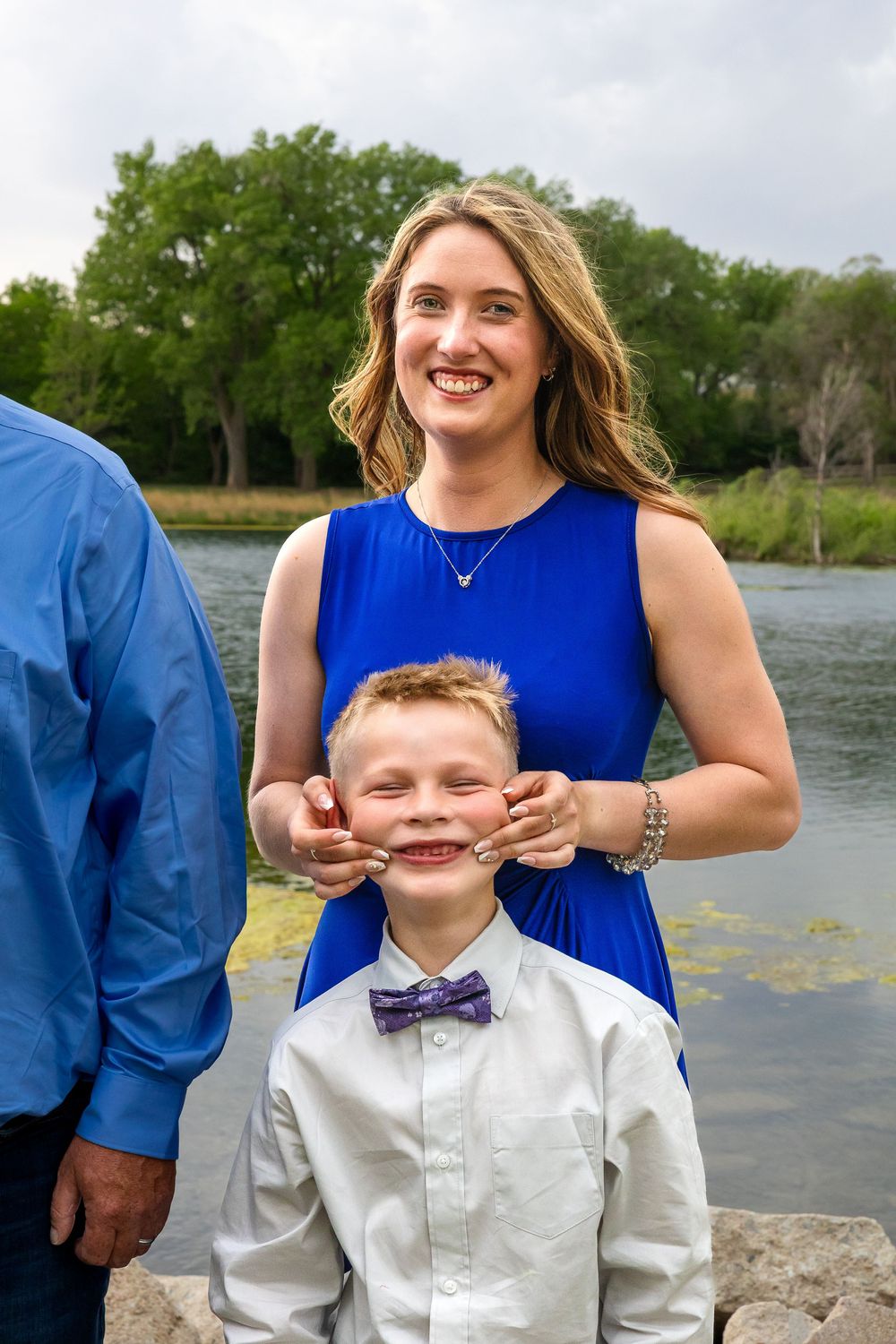 Woman in blue dress playfully stretching boy's cheeks near a river, both smiling during a family photo session.