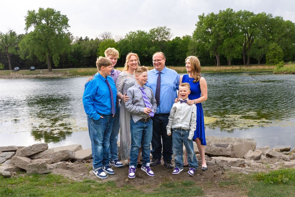 Family of seven posing outdoors near a scenic pond surrounded by lush green trees on a cloudy day.