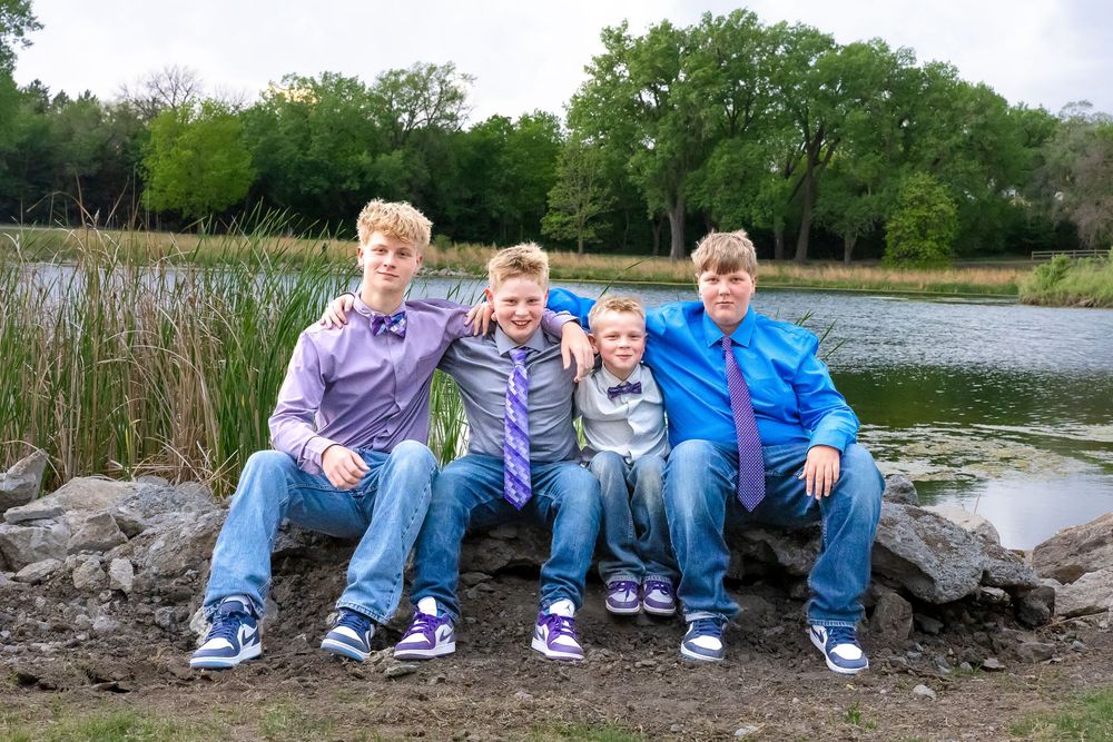 Four boys in dress shirts and jeans sit together on rocks near a pond surrounded by green trees.