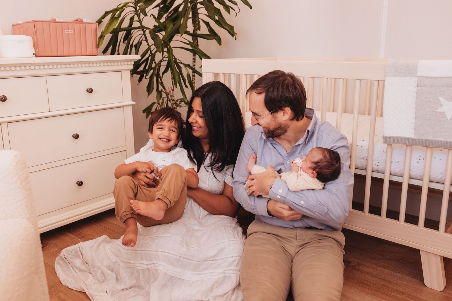 Family of four sits on nursery floor, parents smiling at toddler and newborn baby near white crib and dresser with newborn photographer