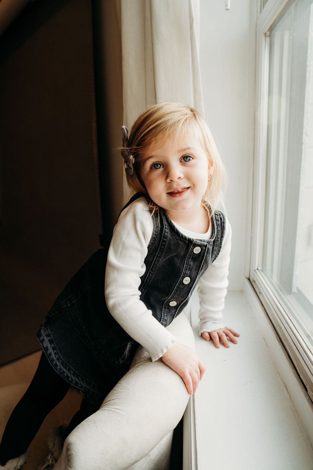 Young blonde girl in denim dress and white top smiles while leaning on a window sill indoors.