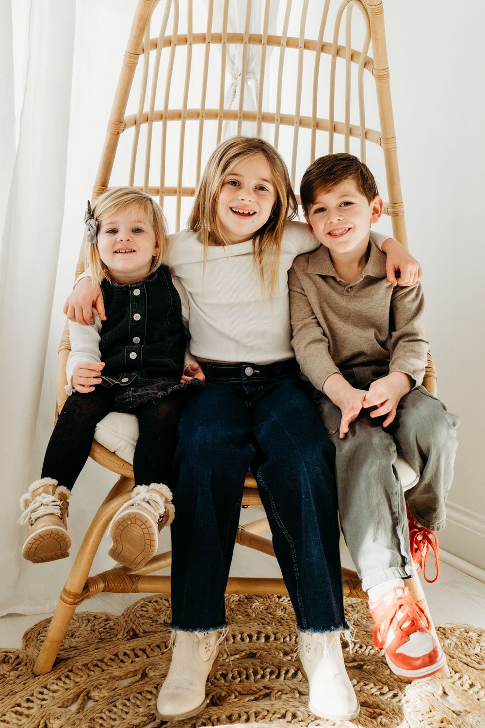 Three smiling children sitting together in a rattan egg chair, dressed in casual outfits for a bright studio photo.