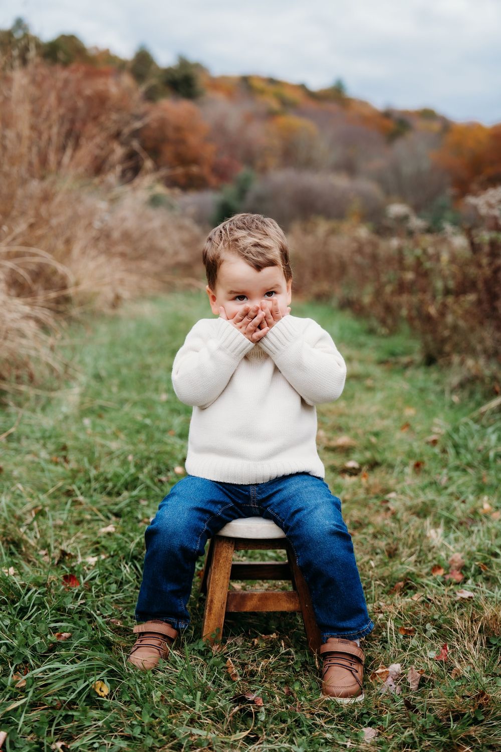 A young boy in a white sweater and jeans sits on a wooden stool outdoors, covering his mouth with both hands.