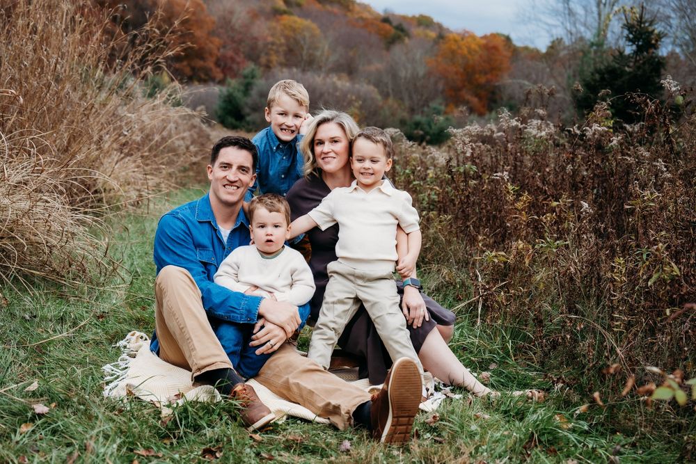 Happy family of five posing outdoors in autumn, sitting on grass surrounded by colorful fall foliage.