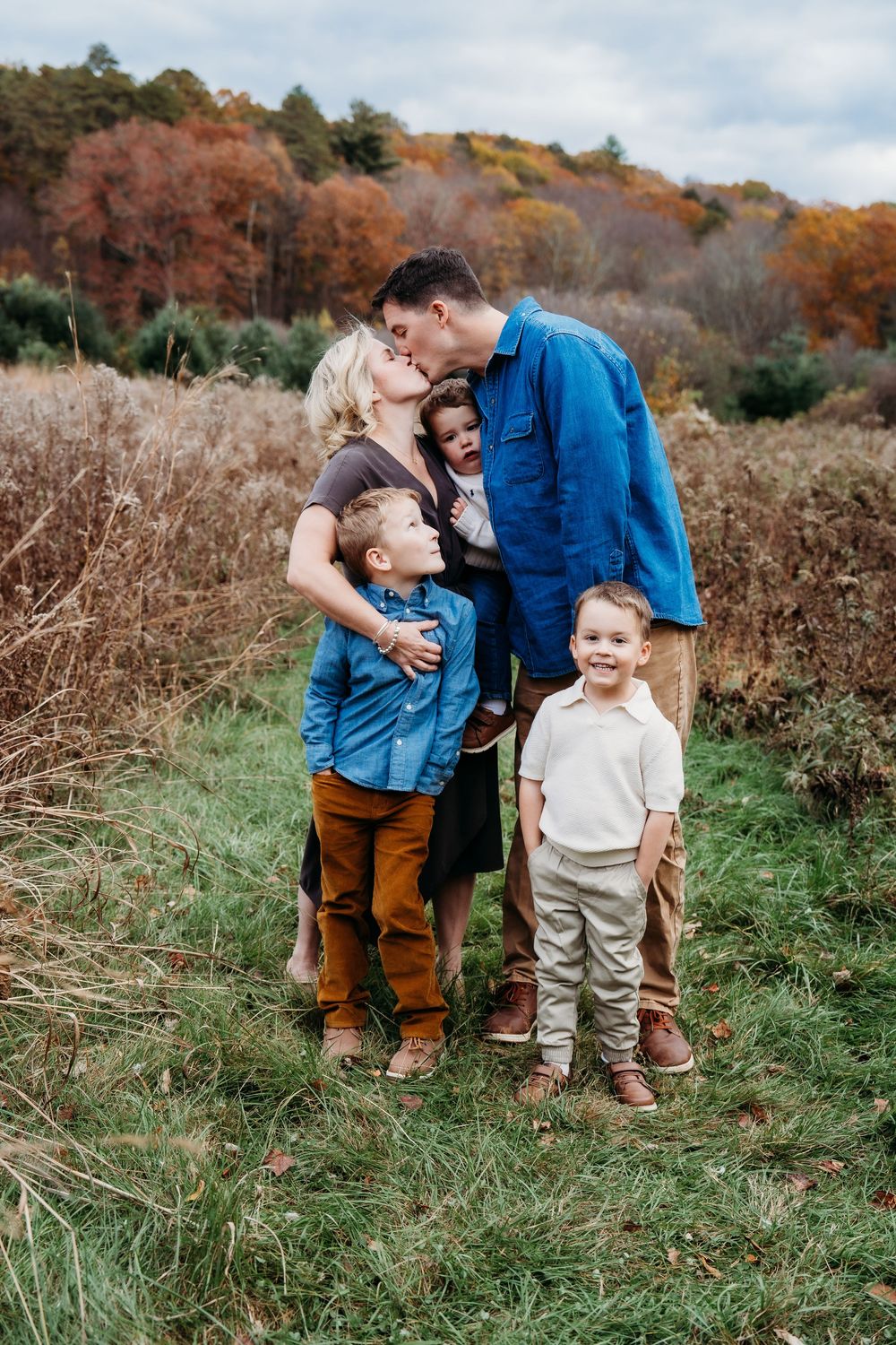 Family of four poses in autumn field, parents kissing child while toddler smiles, colorful fall foliage backdrop.