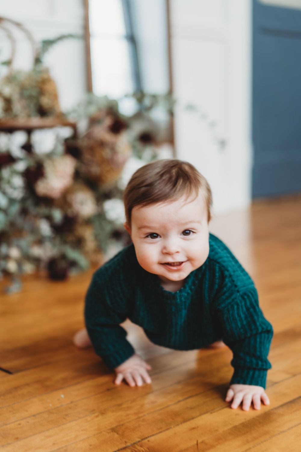 A smiling baby in a green knit sweater crawls on a hardwood floor with blurred plants in the background.