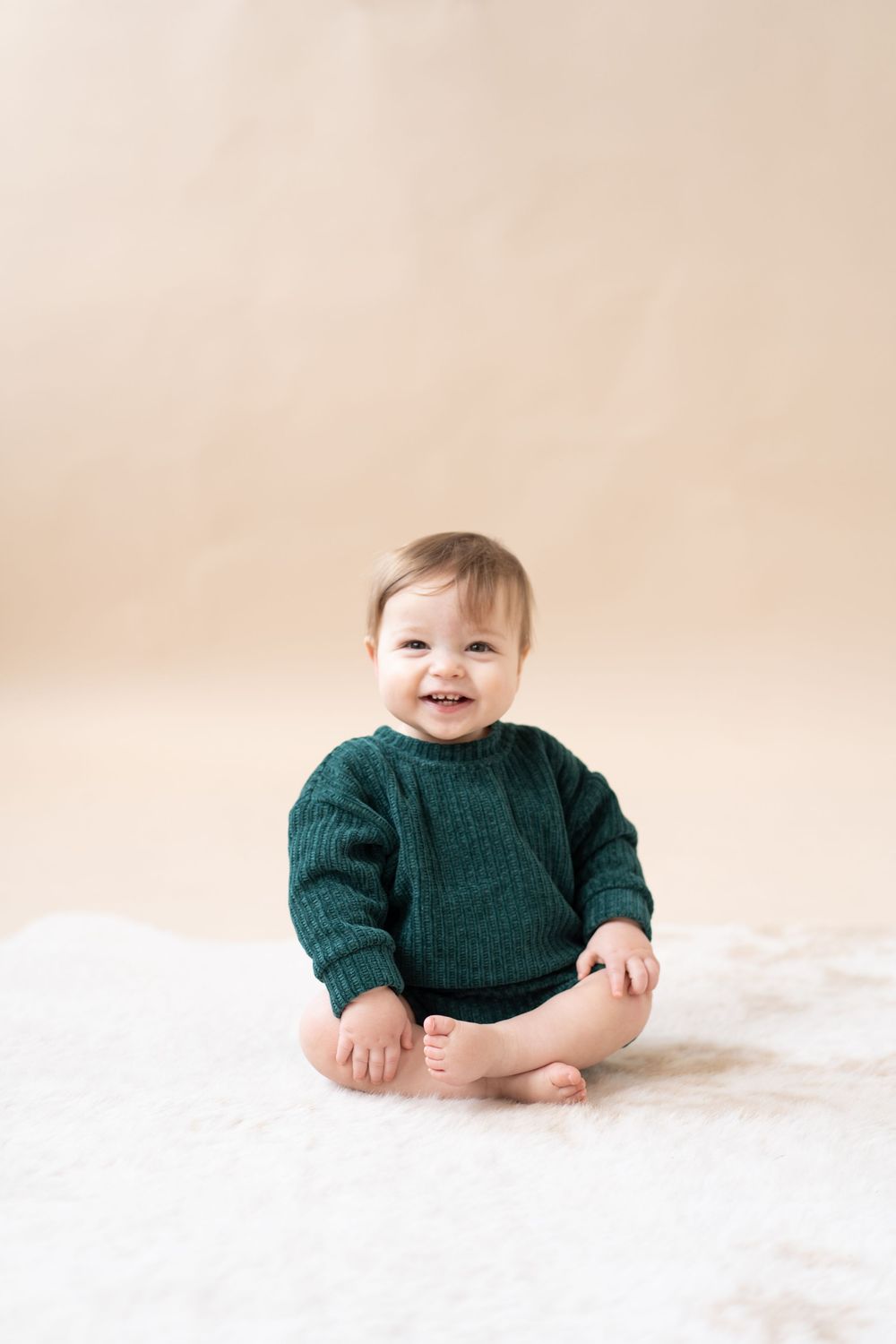 Smiling baby in green knit sweater sitting on white fluffy rug against beige background.