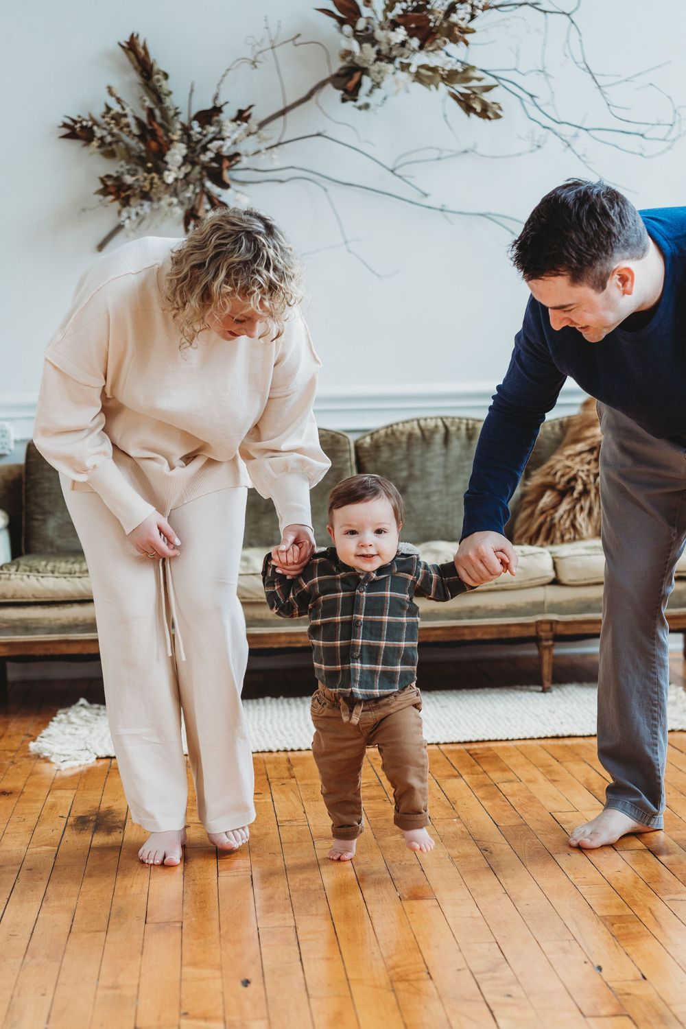 Parents helping their baby take first steps on hardwood floor in cozy living room.