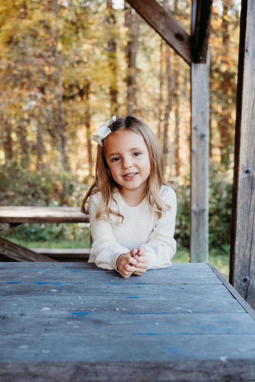 A smiling young girl with a white bow in her hair leans on a wooden table on a rustic porch surrounded by autumn trees.