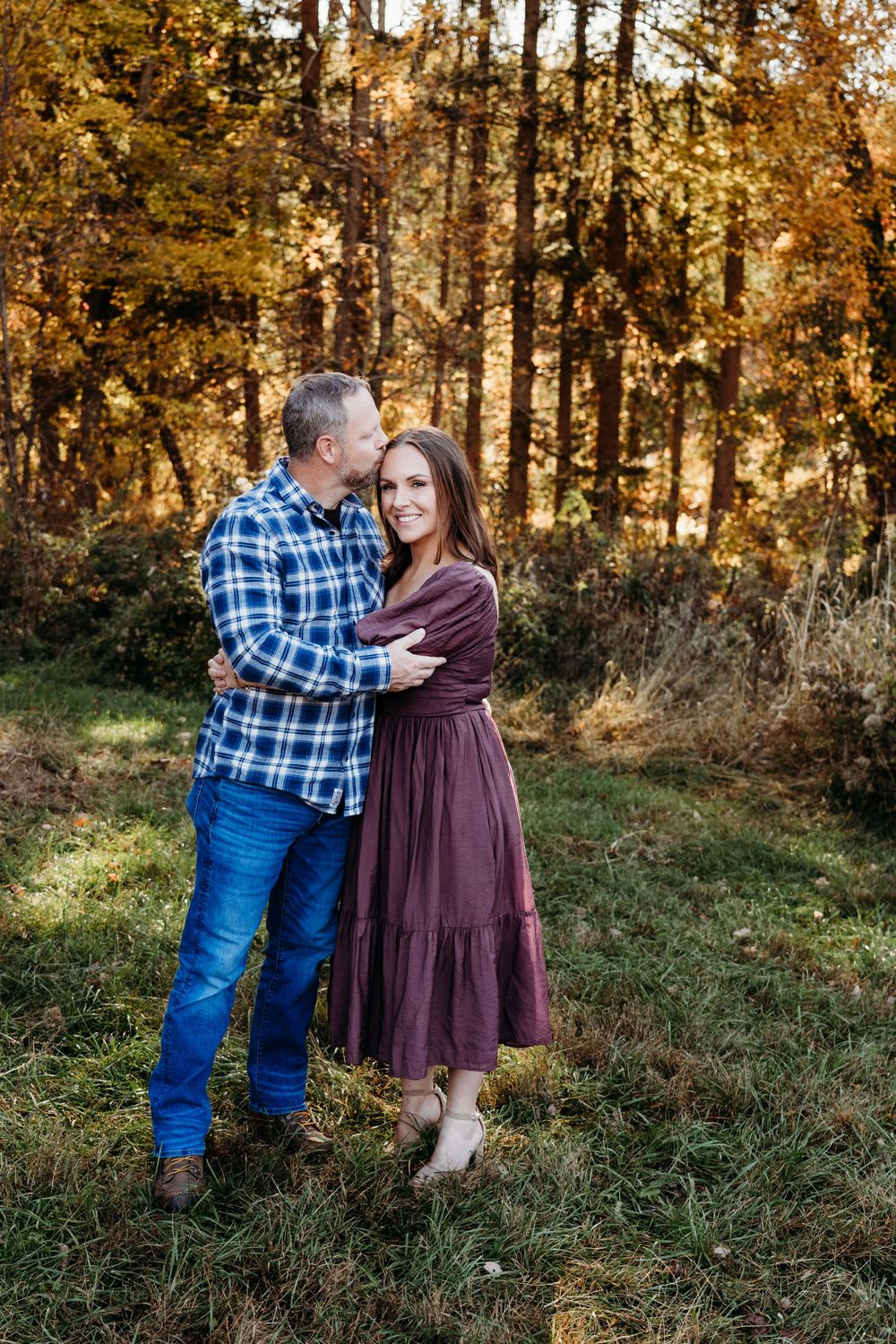 Couple embracing in autumn forest, man in plaid shirt and jeans, woman in burgundy dress, smiling outdoors.