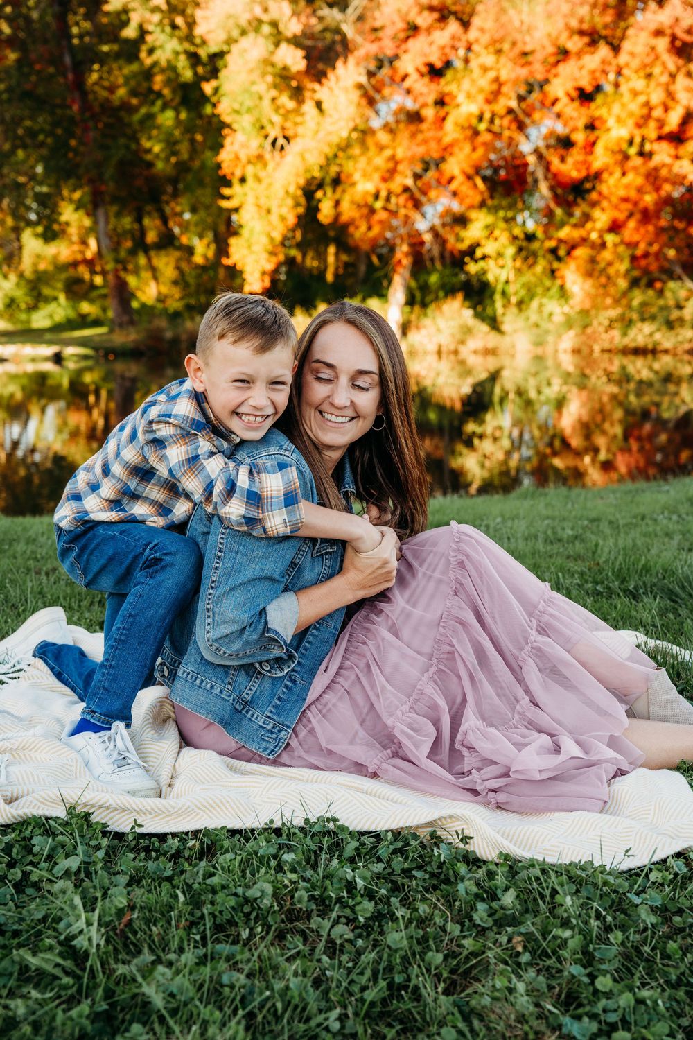 A joyful boy hugs his smiling mother on a blanket outdoors, surrounded by vibrant autumn foliage near a pond.