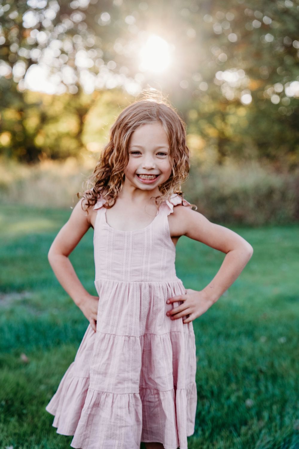 A young girl in a pink tiered sundress smiles with hands on hips in a sunny green outdoor setting.