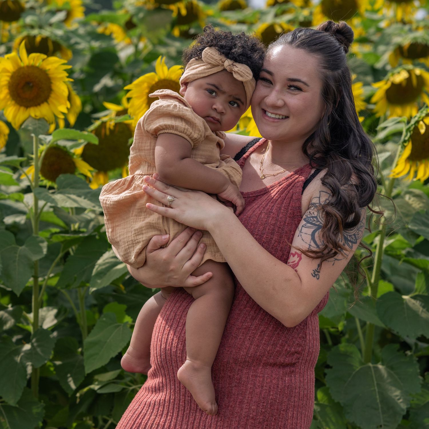 A smiling young woman holds a baby in a sunflower field for New Hampshire sunflower mini sessions