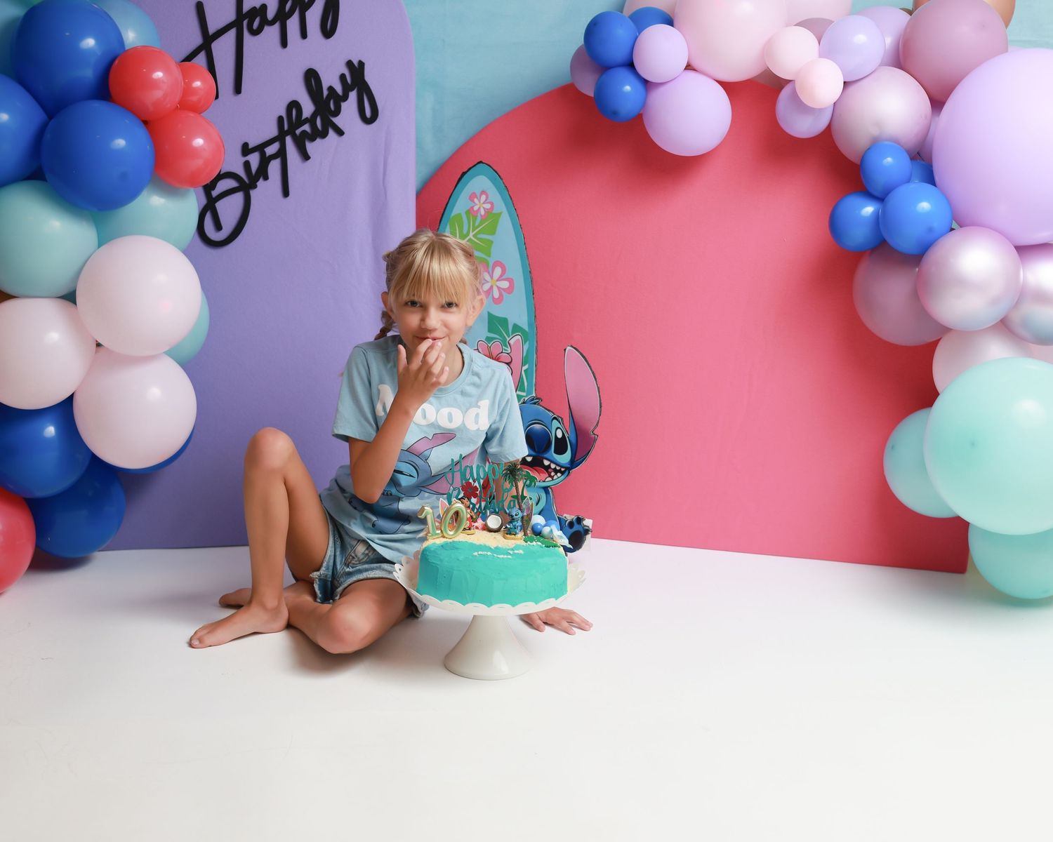 Child sitting on floor with Lilo & Stitch birthday cake, surrounded by colorful balloon arch and Happy Birthday backdrop.
