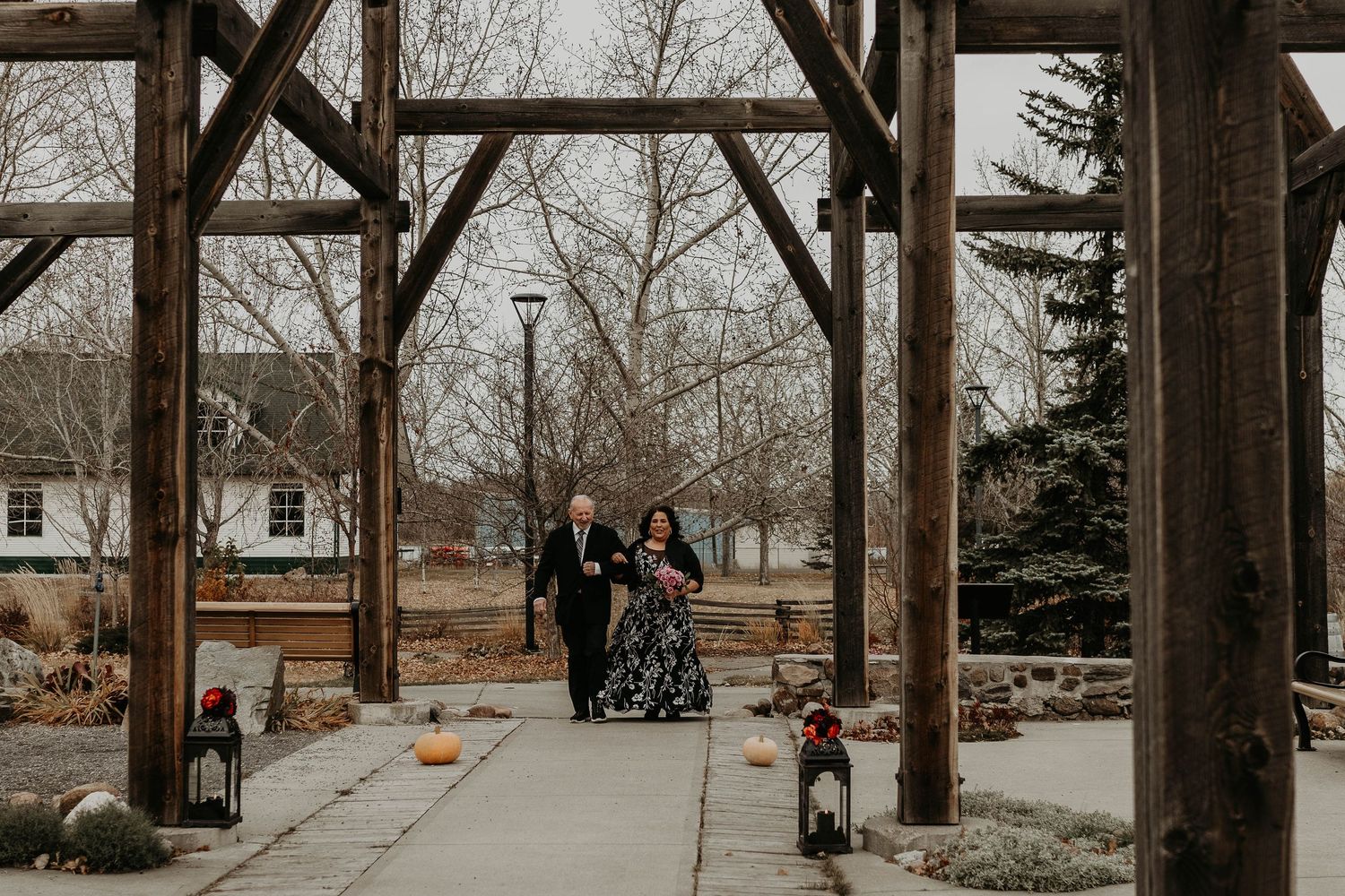 Couple walks under rustic wooden pergola at outdoor wedding venue with bare trees and pumpkin decorations.