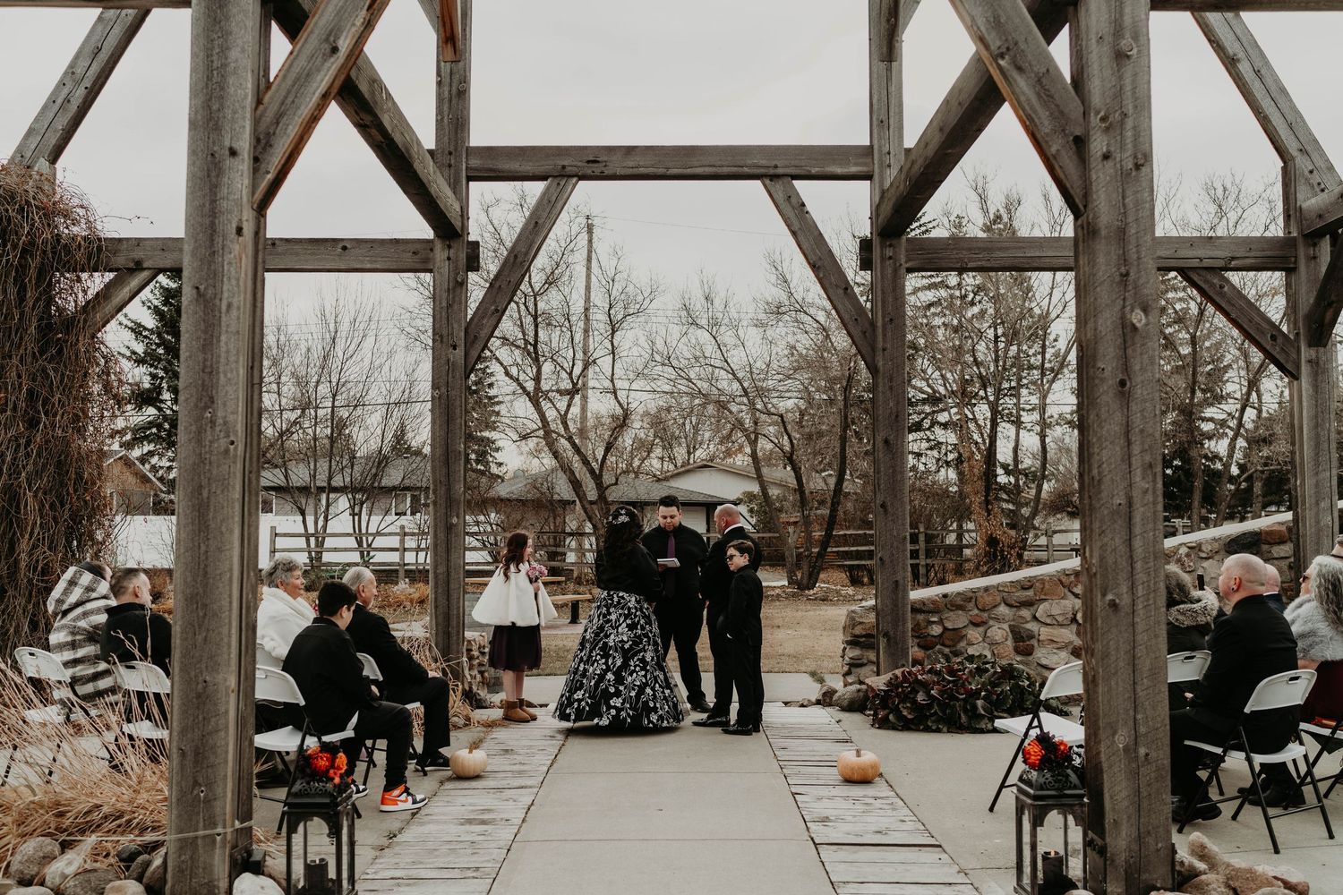 Outdoor winter wedding ceremony under rustic wooden pergola with guests seated on black chairs lining the aisle.