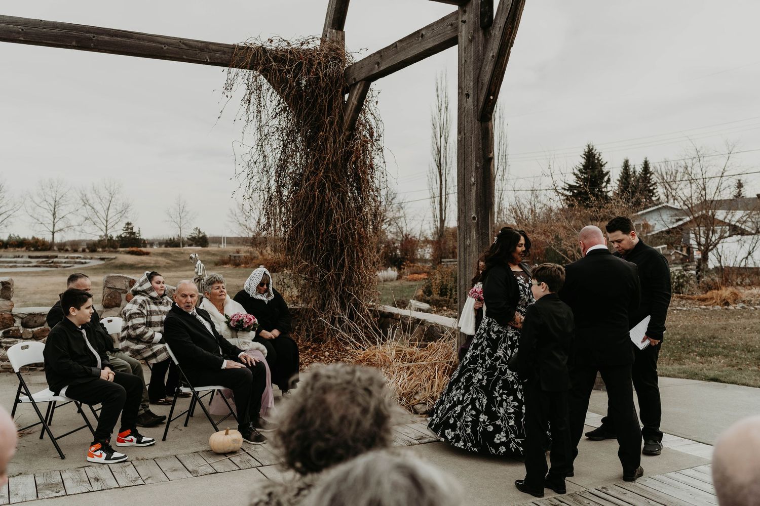 Outdoor wedding ceremony under a rustic pergola with guests seated on chairs in a natural garden setting.