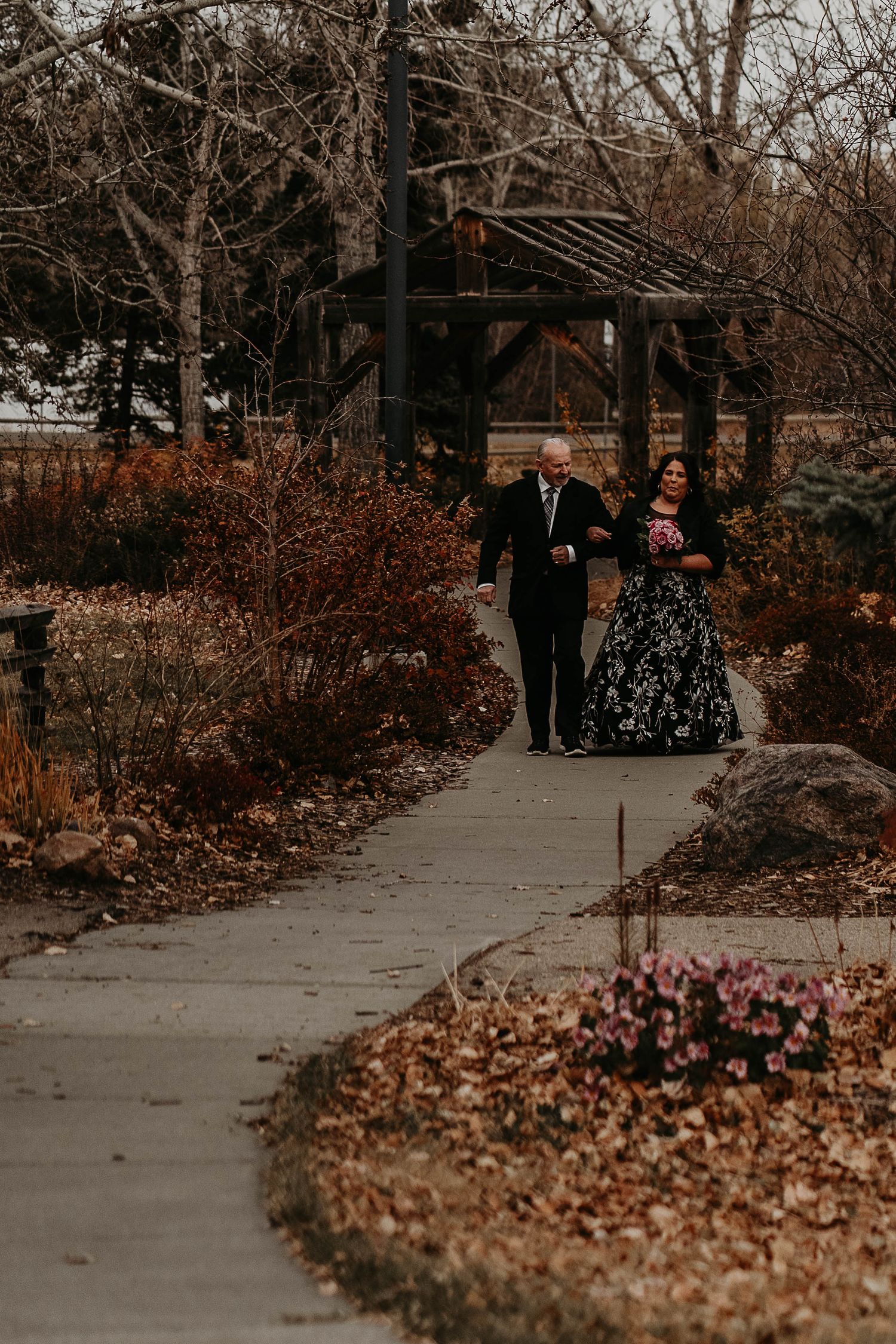 Bride in black floral gown walks with escort along garden path amid autumn foliage near rustic wooden pergola.