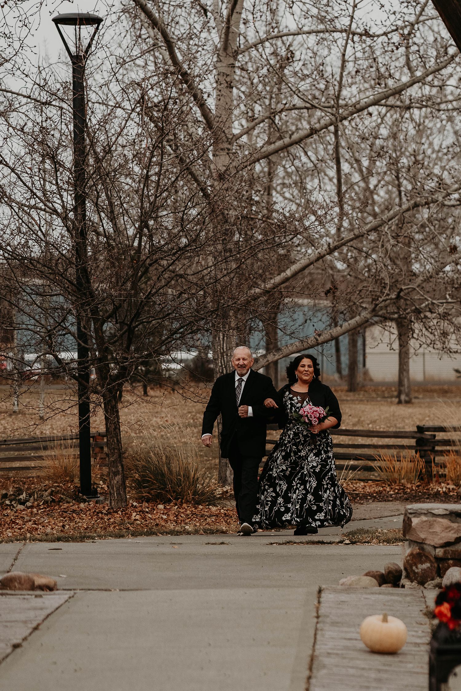 Couple walks along a park path in autumn, woman in floral gown holding pink bouquet, bare trees and lamp post behind them.