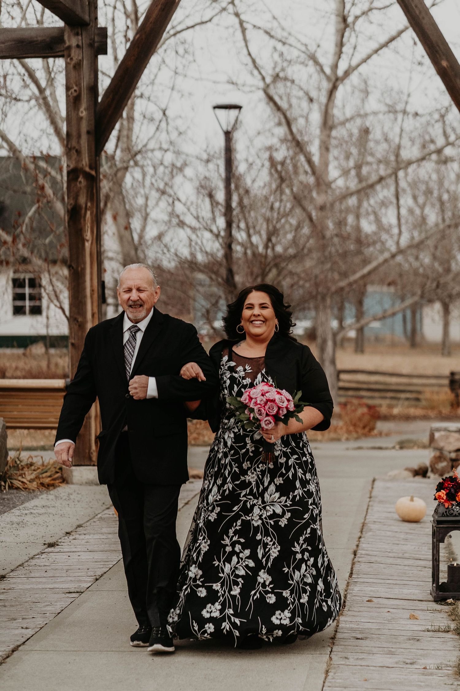 Couple walks arm-in-arm outdoors, woman in black floral gown holding pink bouquet, man in black suit, at a winter wedding.