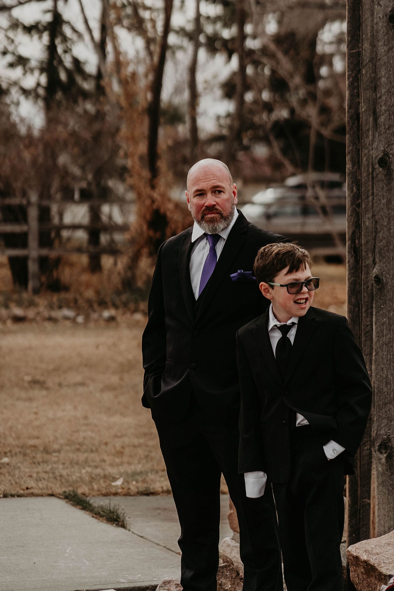 A bald man in a black suit with purple tie stands beside a boy in sunglasses and suit at an outdoor wedding.