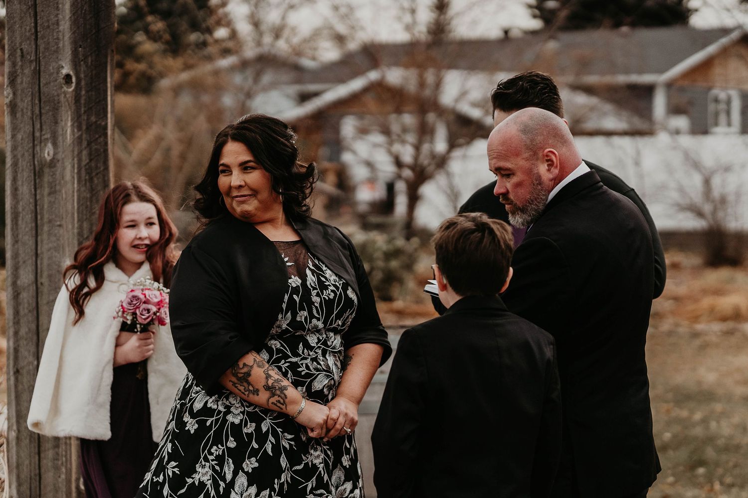 Outdoor wedding ceremony with bride in black floral dress, groom in suit, and children attendants in winter setting.
