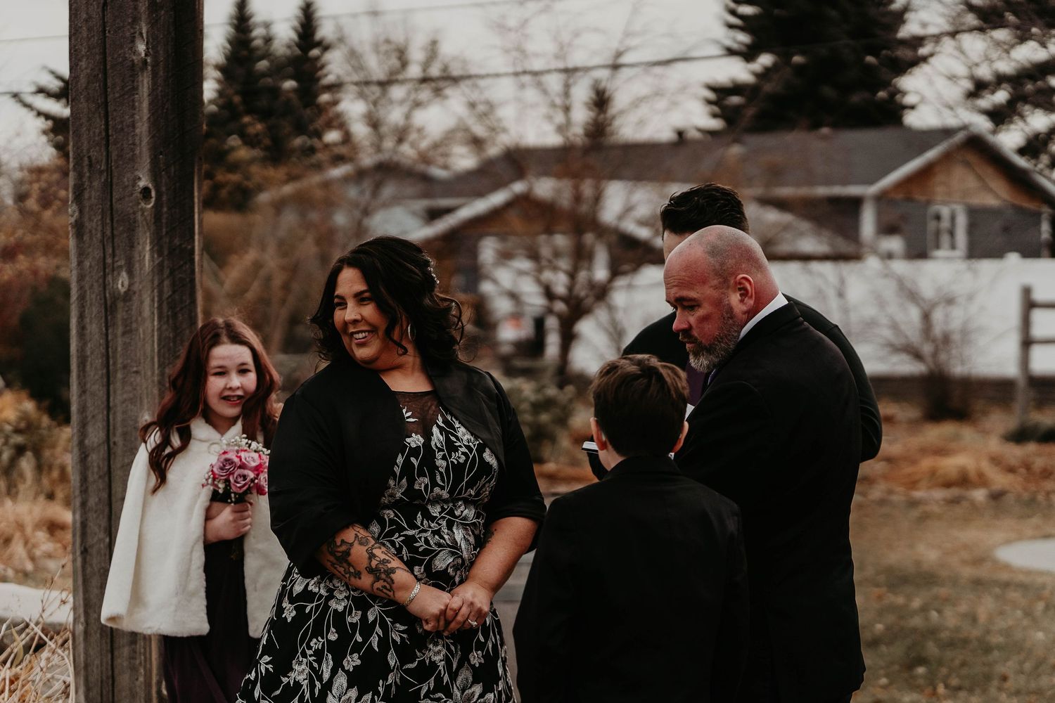 Outdoor wedding ceremony with bride in floral dress, groom in suit, and children attendants, set against a rustic backdrop.