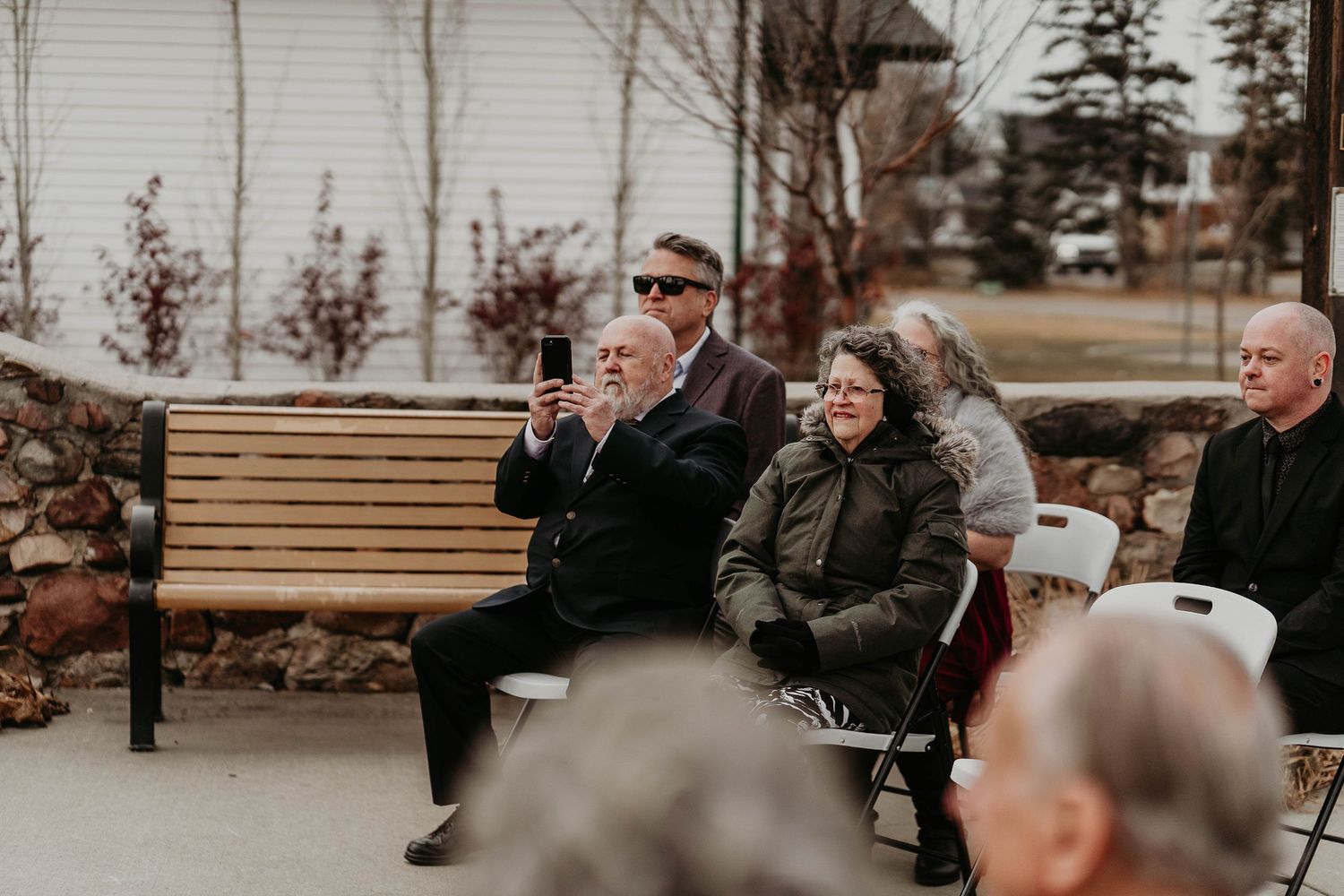 Guests seated outdoors at a wedding ceremony, with one man taking a photo on his phone on a cloudy day.