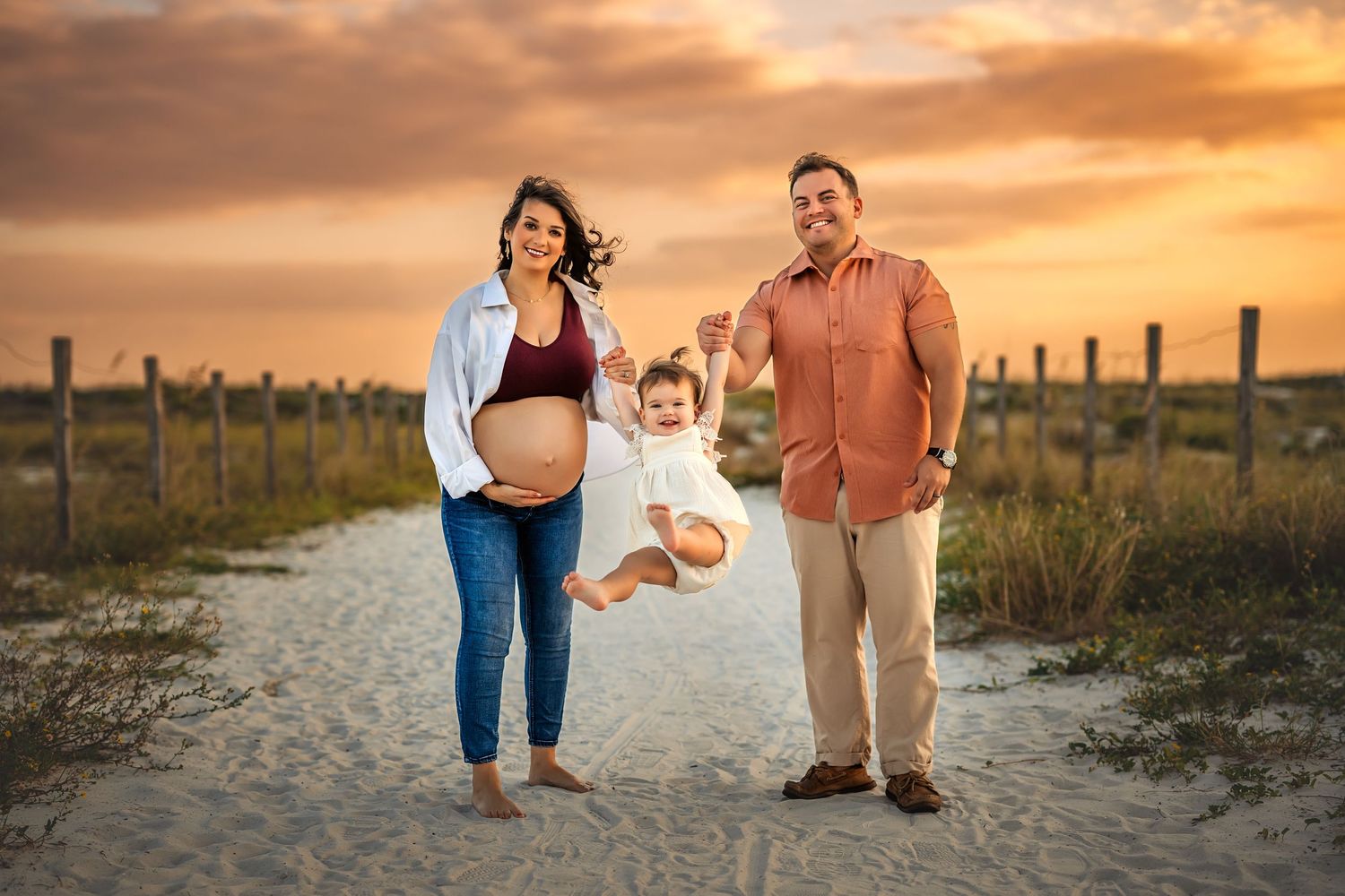 Pregnant woman and man swing toddler between them on sandy beach path at golden sunset.
