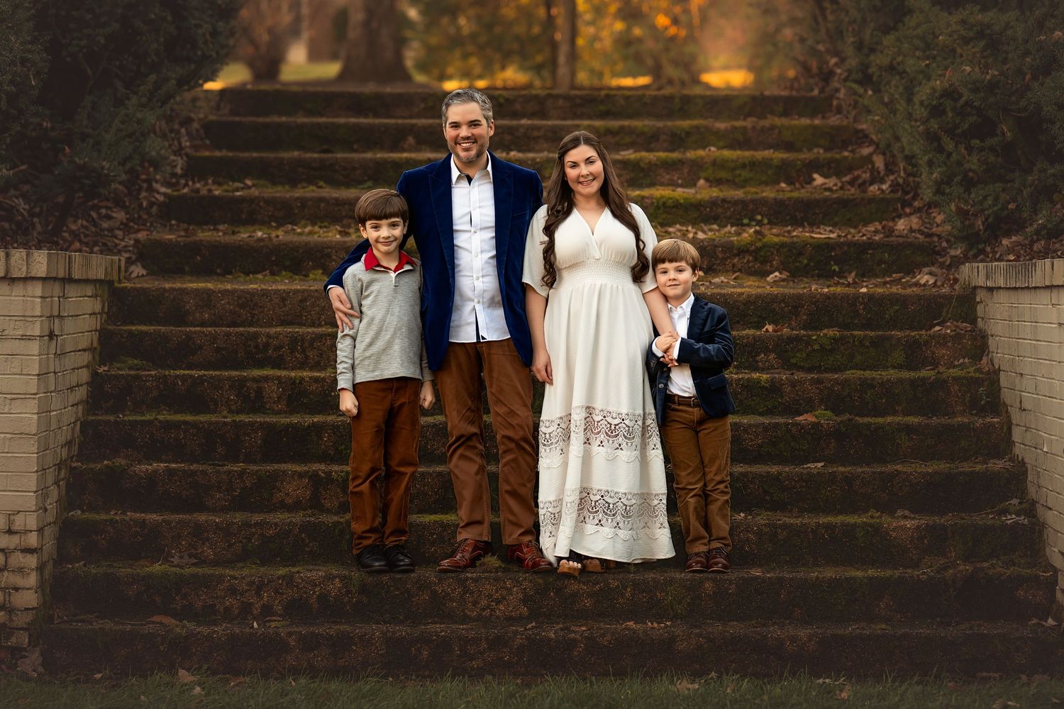 Family of four poses on stone steps surrounded by autumn foliage, mother in white dress, father and two young sons smiling.