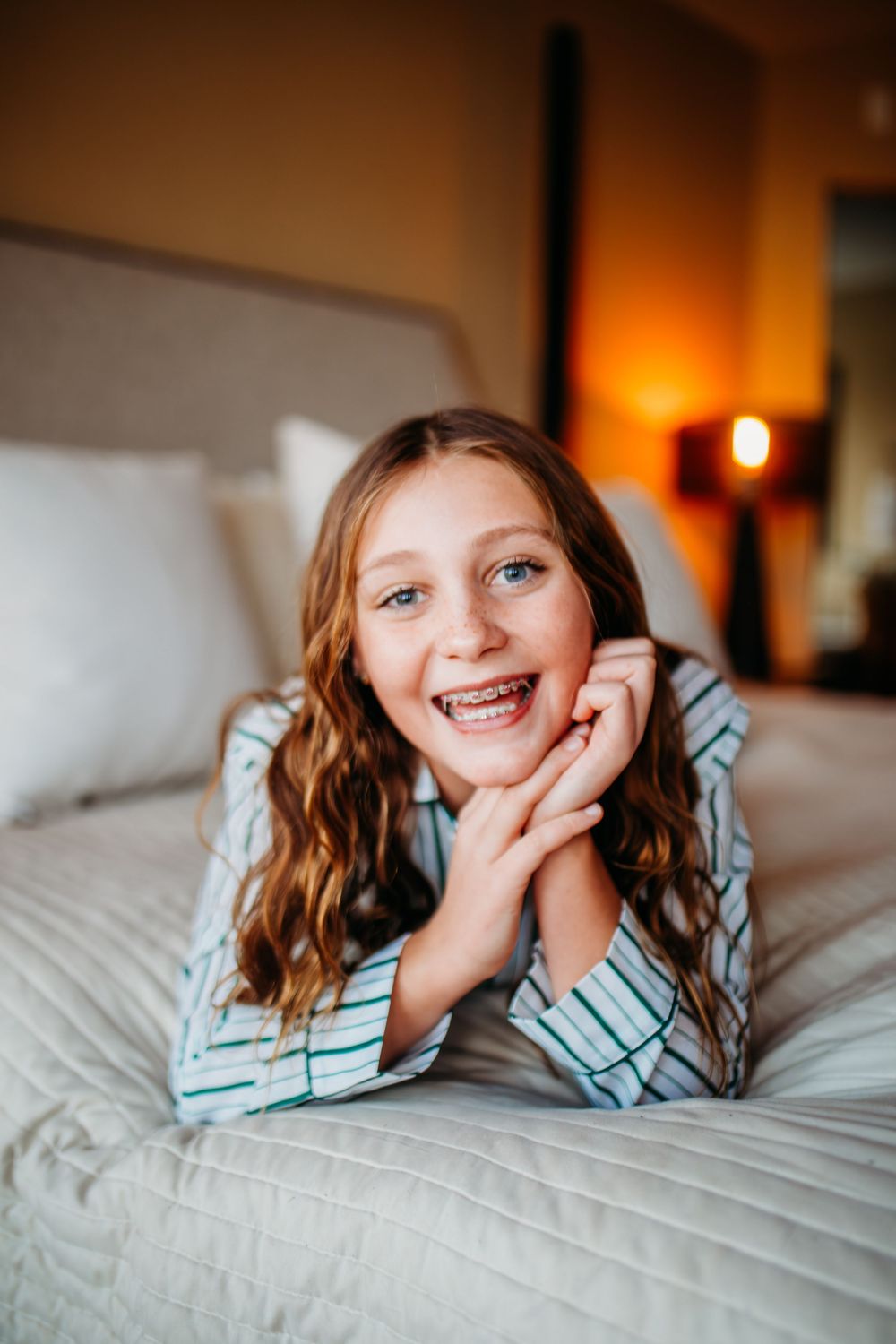 Smiling girl with braces and wavy hair lying on a bed in striped pajamas.