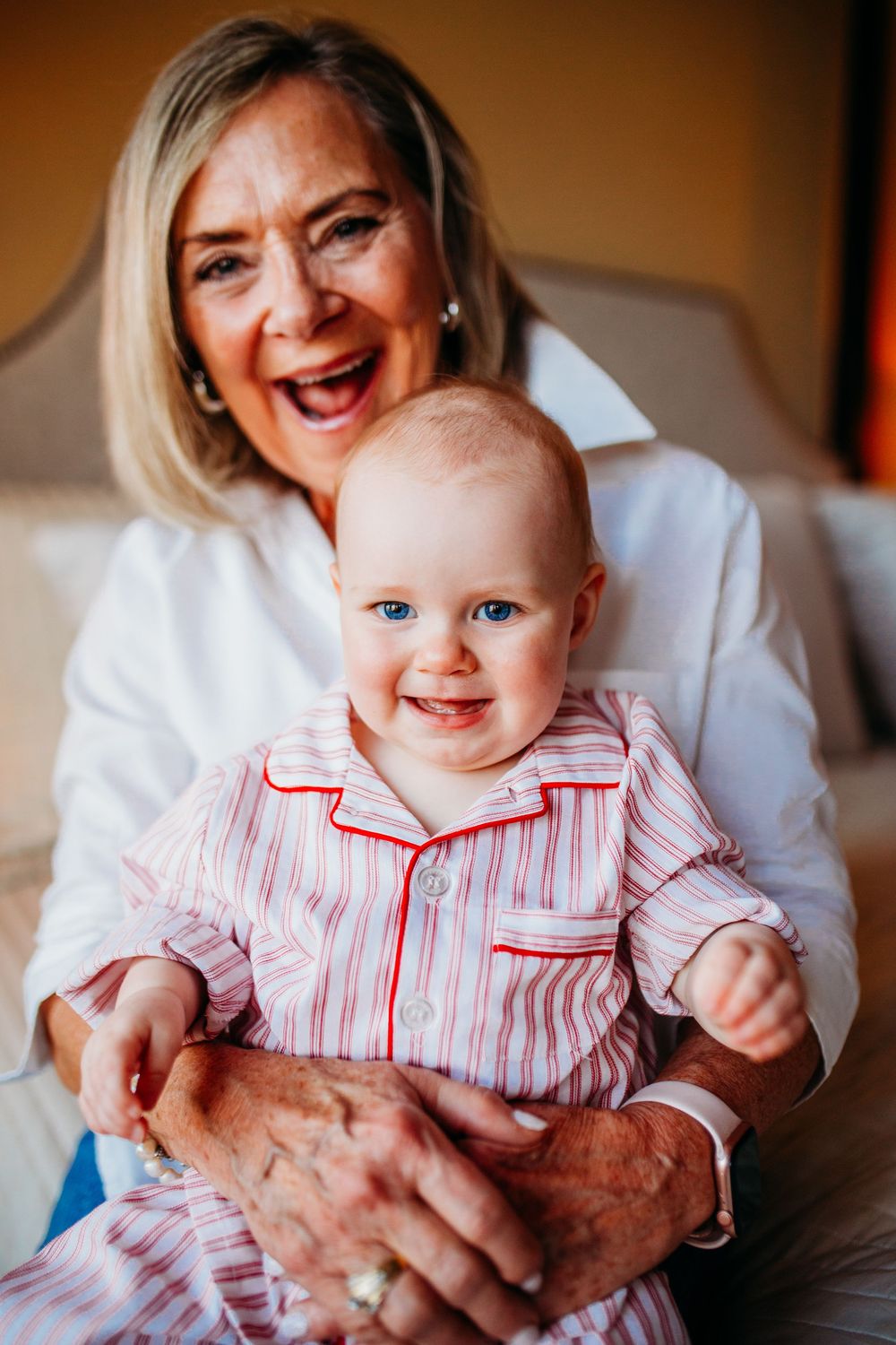 Elderly grandmother joyfully holding smiling baby in pink striped pajamas, both looking at camera.
