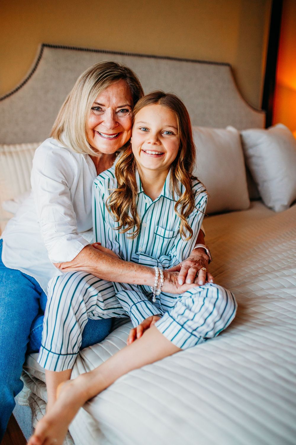 A smiling woman and young girl in striped pajamas sitting together on a white bed in a cozy bedroom.