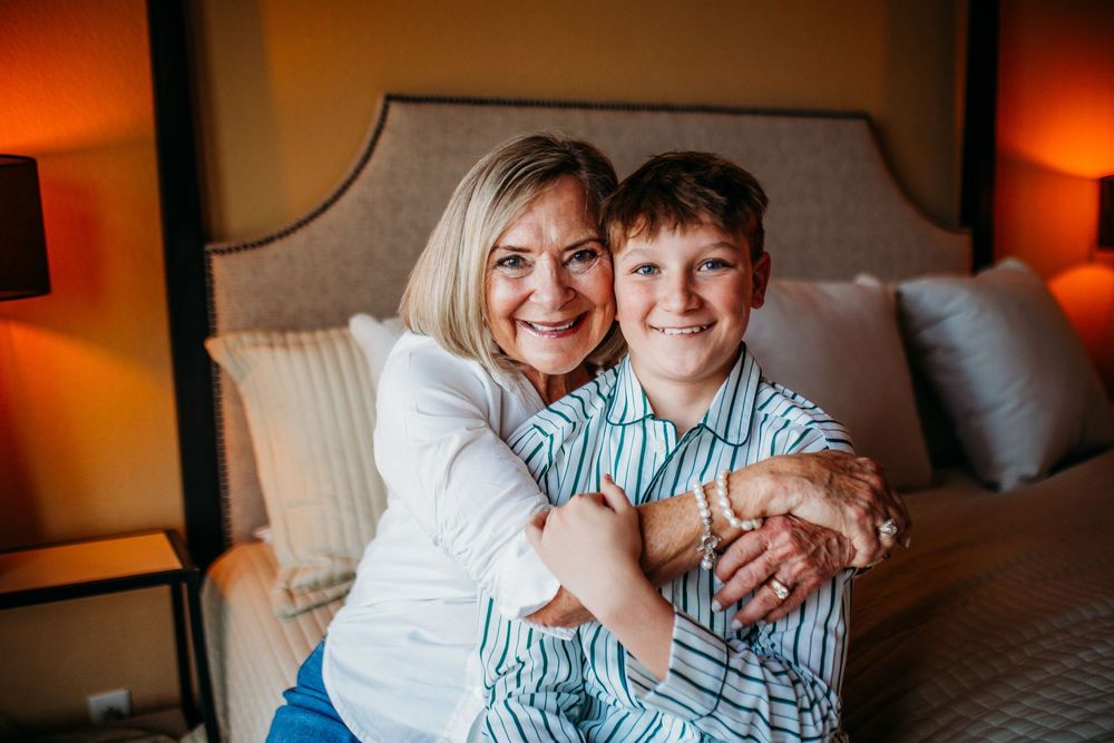 A woman and young boy share a warm hug while sitting in front of a bed in a cozy hotel room.